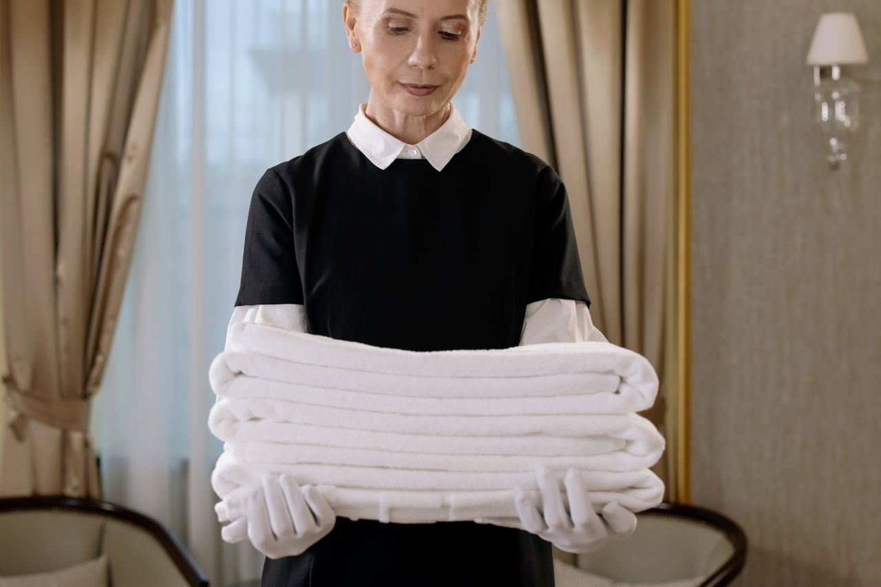 services-03 Professionnel de l’entretien tenant une pile de serviettes fraîches dans une chambre d’hôtel luxueuse, illustrant élégance et qualité de service.